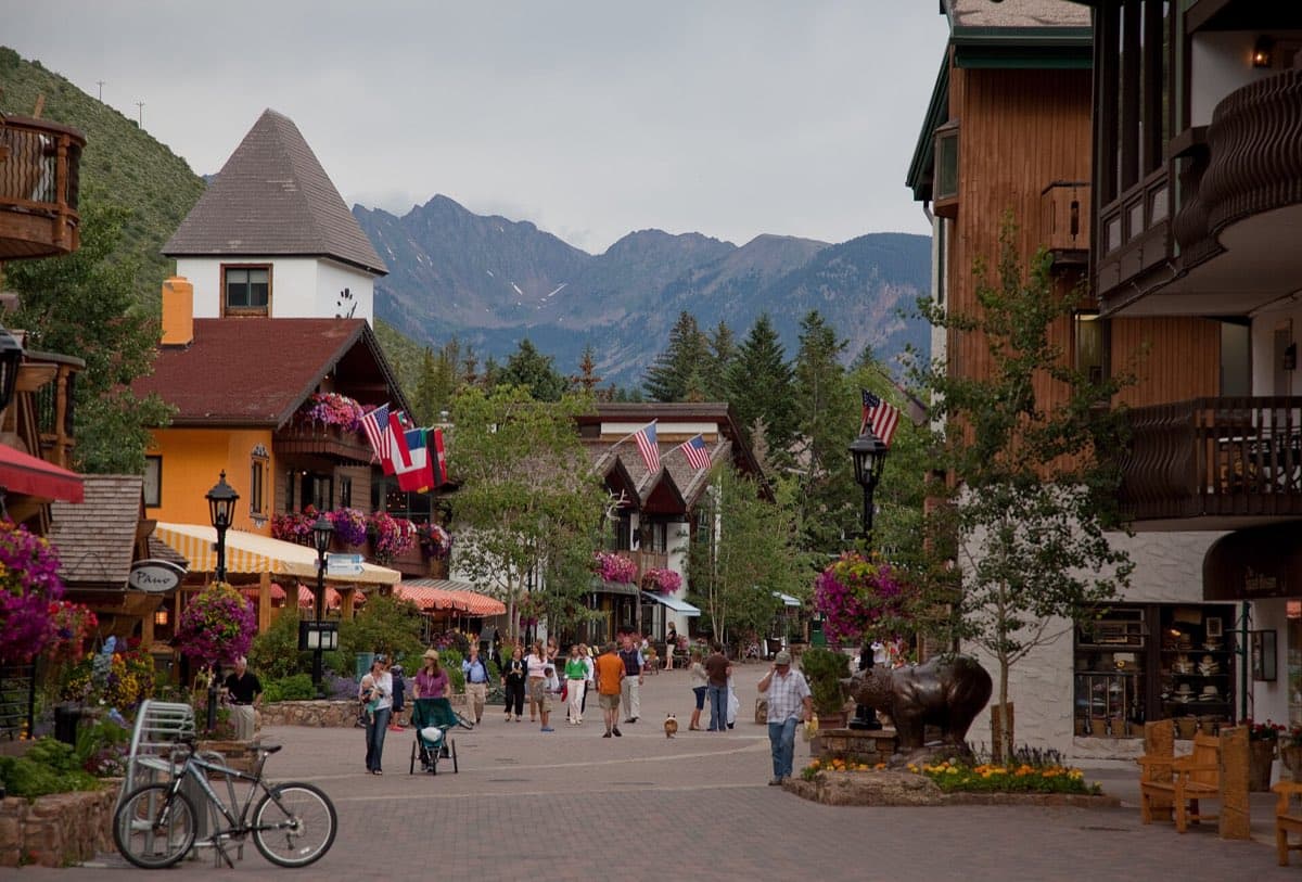 Pedestrians and flower-filled buildings along Gore Creek Drive in Vail, with the jagged Gore Range rising behind the village.
