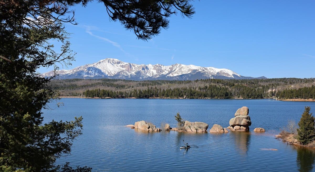 A kayaker crossing blue Rampart Reservoir beneath granite boulders and the snow-covered north face of Pikes Peak.