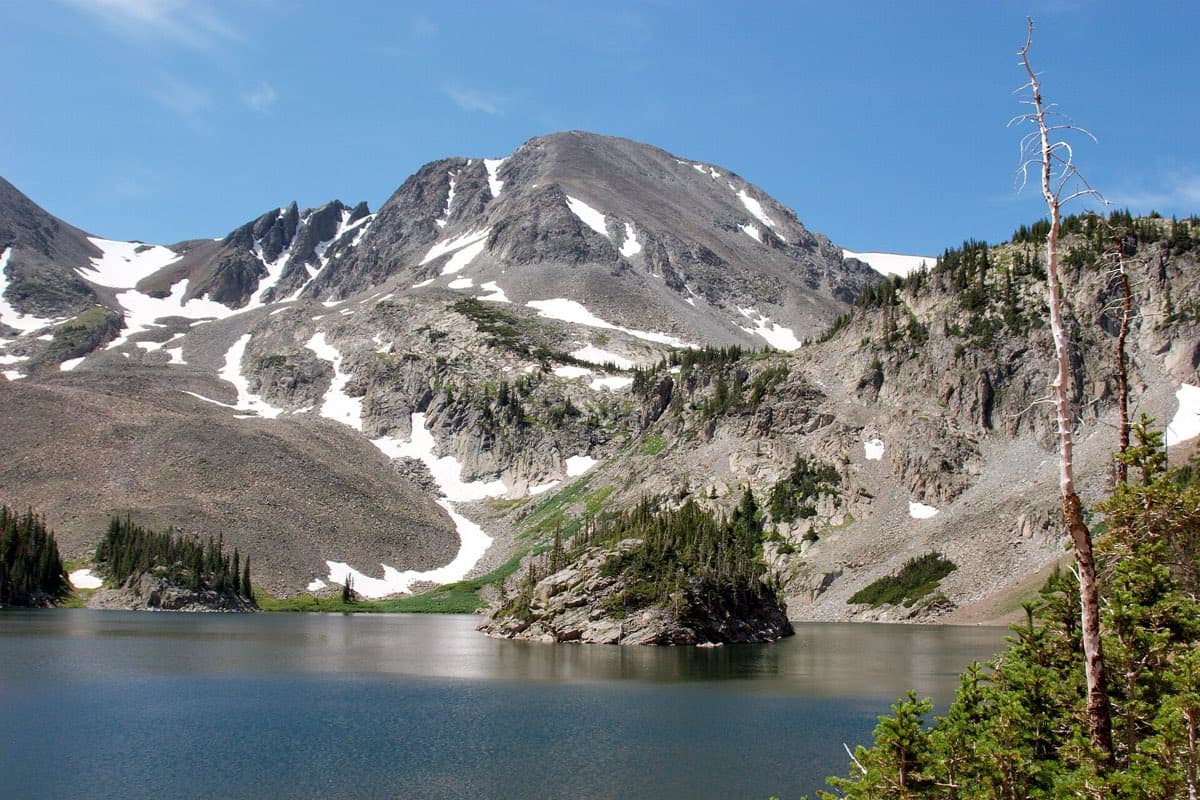 Rocky alpine slopes with lingering snow rise above Lake Agnes and a small tree-covered island in State Forest State Park.