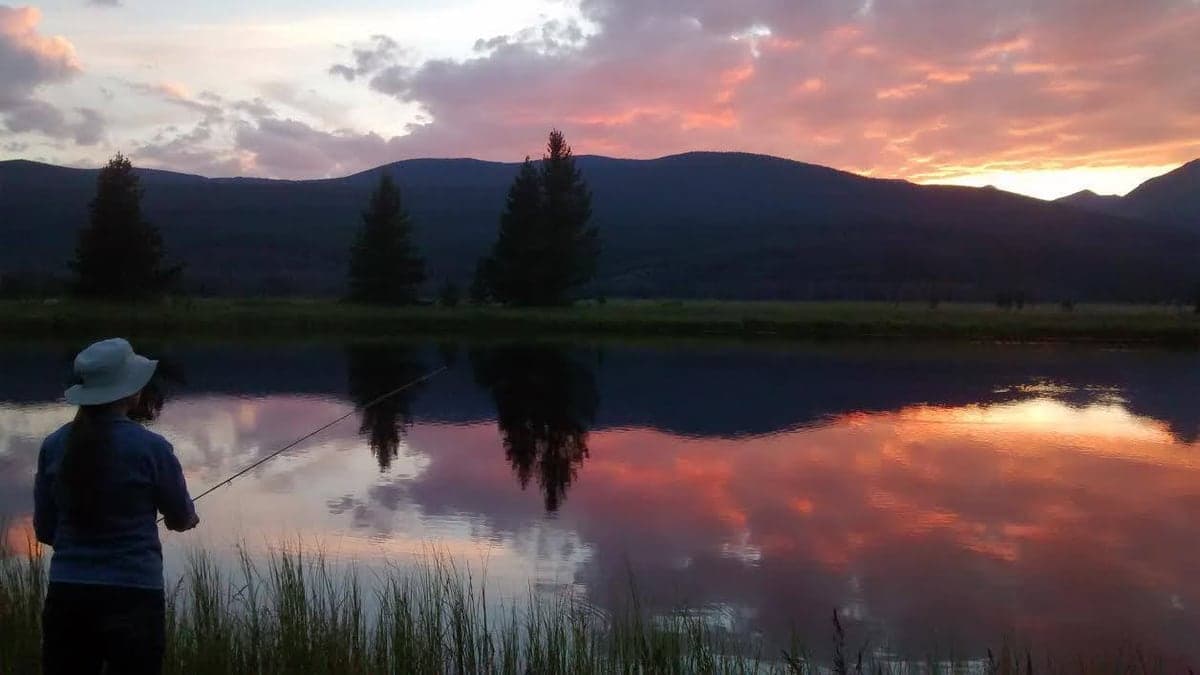 A person fishing in Kawuneeche Valley as a pink sunset reflects below the Never Summer Range