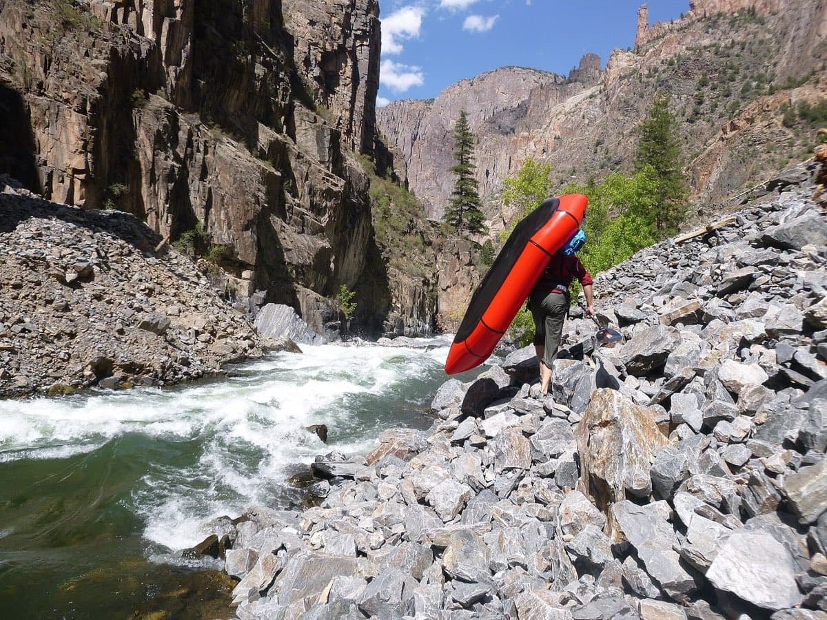 A rafter carries a red packraft beside whitewater and steep rock walls on the Gunnison River route from East Portal.