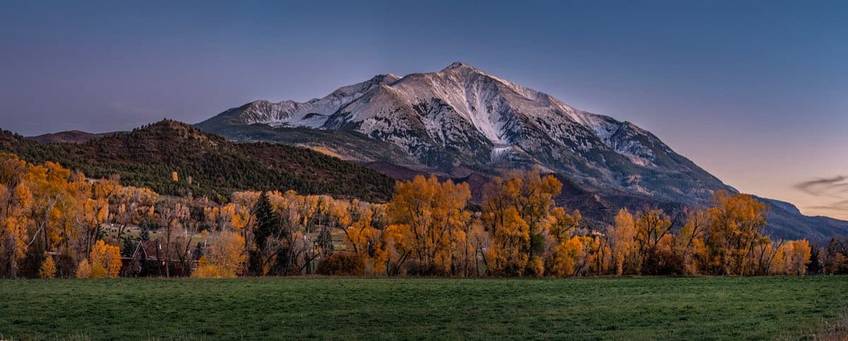Snow-dusted Mount Sopris rises above golden autumn cottonwoods and a green field near Carbondale at evening.