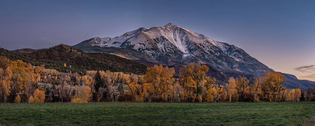 Snow-dusted Mount Sopris rises above golden autumn cottonwoods and a green field near Carbondale at evening.